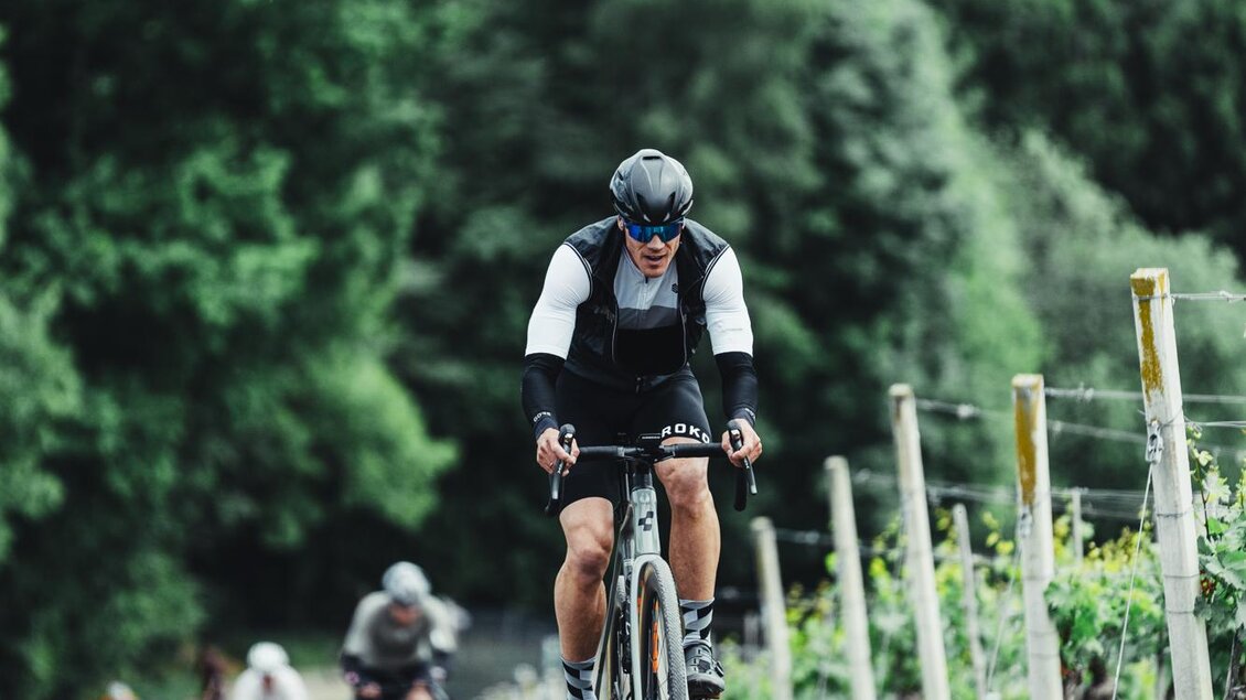 Ein Radfahrer fährt einen mit Kies bedeckten Weg bergauf, umgeben von Weinreben. Im Hintergrund sind grüne Hügel und Bäume sichtbar. | © hofstaetter@finisher-magazin.at