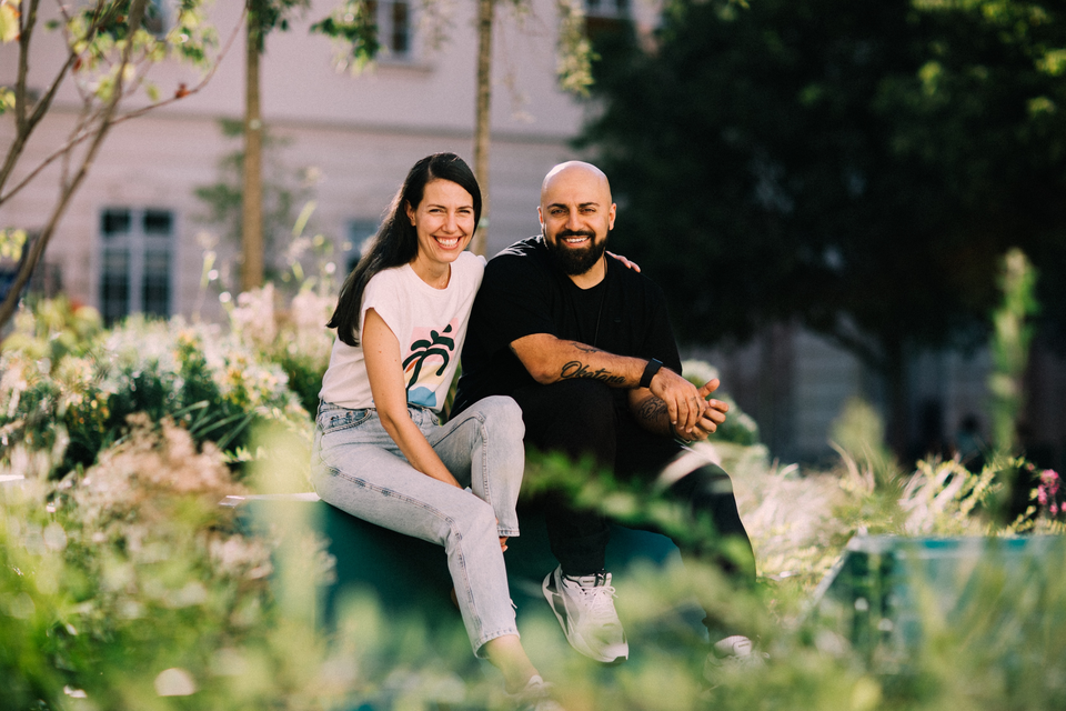 Zwei Personen sitzen im Grünen und lächeln in einem Park in Graz, umgeben von Pflanzen und Bäumen. | © Ali Mahlodji