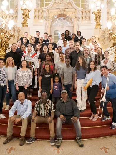 A large group of people is posing on a magnificent staircase. The room is elegantly designed with golden embellishments and intricate details.