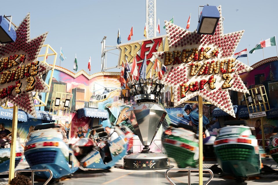 Ein lebhaftes Volksfest mit bunten Fahrgeschäften und vielen Fahnen. Menschen haben Spaß auf den Fahrten inmitten einer festlichen Atmosphäre. | © Valerie_Maltseva
