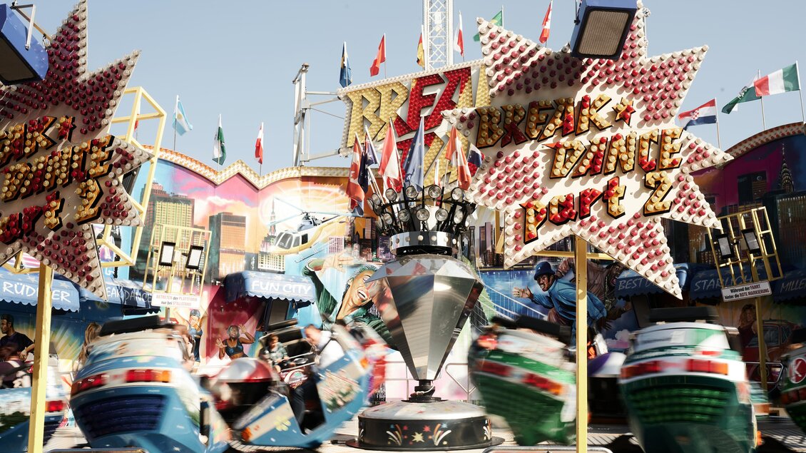 Ein lebhaftes Volksfest mit bunten Fahrgeschäften und vielen Fahnen. Menschen haben Spaß auf den Fahrten inmitten einer festlichen Atmosphäre. | © Valerie_Maltseva