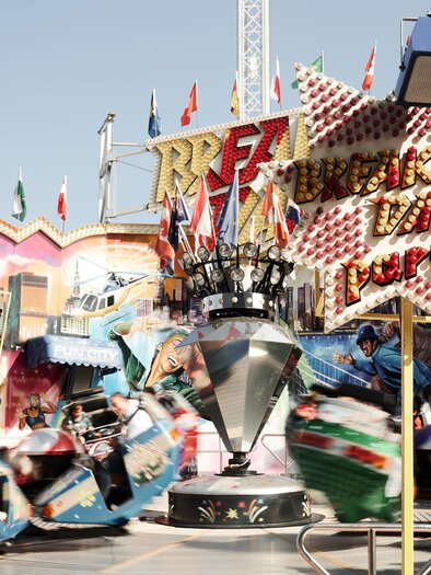 Ein lebhaftes Volksfest mit bunten Fahrgeschäften und vielen Fahnen. Menschen haben Spaß auf den Fahrten inmitten einer festlichen Atmosphäre. | © Valerie_Maltseva