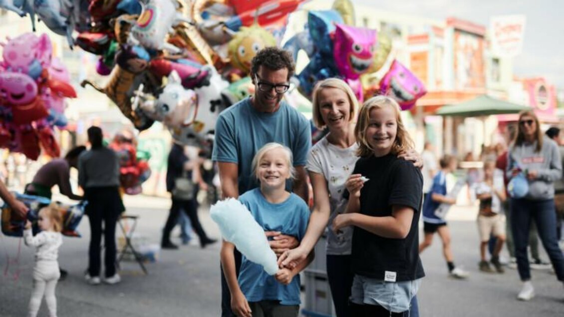 Eine fröhliche Familie steht auf einem Fest mit bunten Luftballons im Hintergrund. Die Kinder halten Zuckerwatte in der Hand und lächeln in die Kamera. | © Valerie_Maltseva