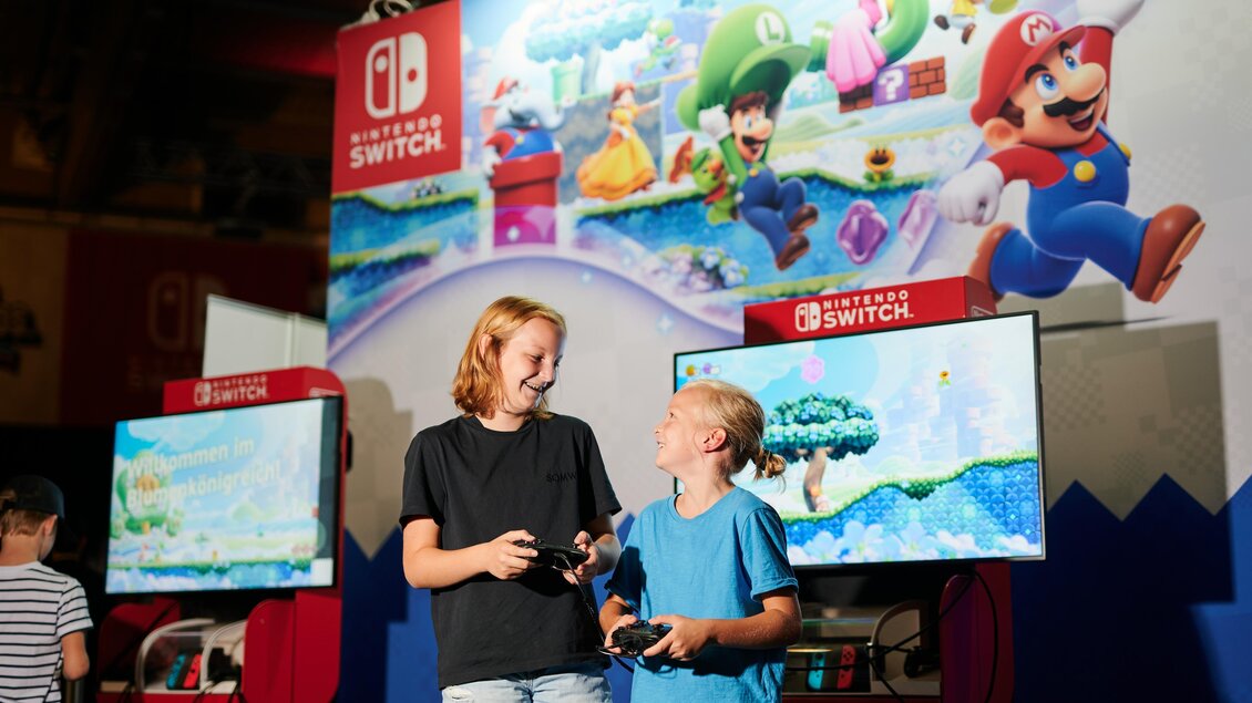 Zwei Kinder spielen zusammen an Nintendo Switch Konsolen. Im Hintergrund ist ein großes Bild von Mario und anderen Charakteren zu sehen. | © Valerie_Maltseva