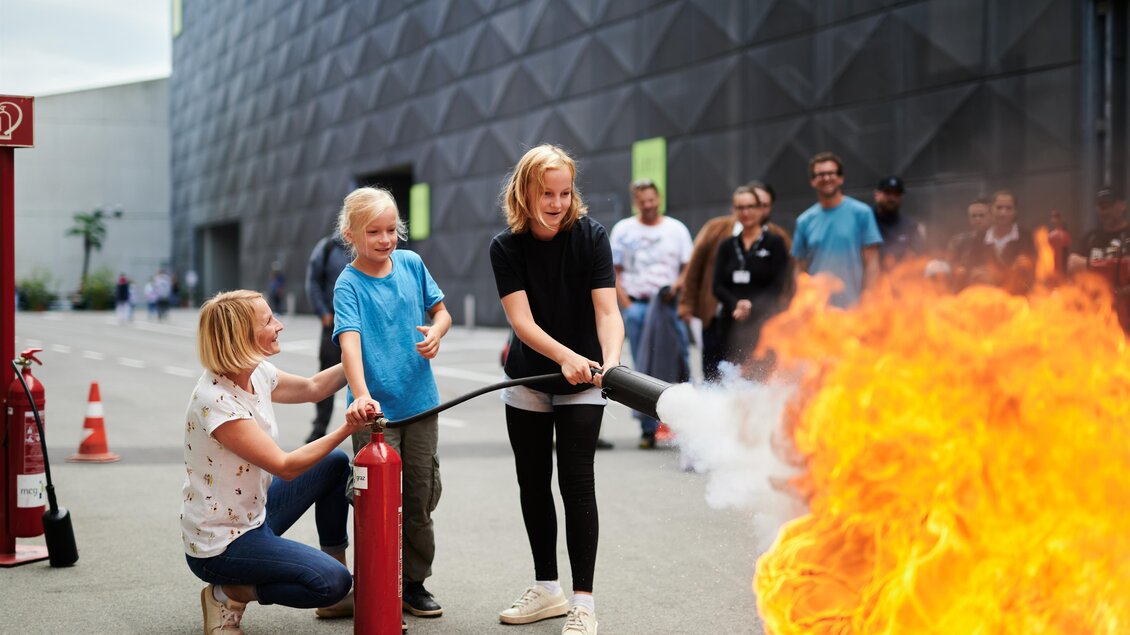 Eine Gruppe von Kindern und Erwachsenen übt den Einsatz eines Feuerlöschers. Im Hintergrund ist ein großes Feuer zu sehen, das simuliert wird. | © Valerie_Maltseva