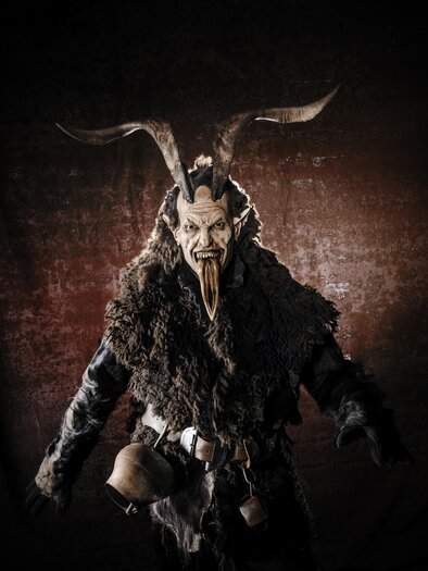 A mysterious man in a costume with a horned head and a terrifying face. He is wearing a fur jacket and is positioned against a dark background. | © © Croce und WIR