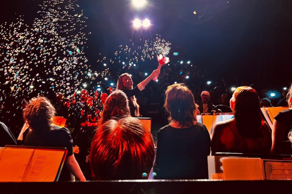 Ein Konzert mit einem Dirigenten, der eine Rose in der Hand hält. Im Hintergrund sind Musiker und eine festliche Atmosphäre mit Lichtern und Seifenblasen zu sehen. | © Georg-Kroneis.