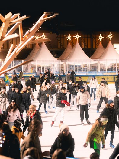 Die belebte Eisfläche der Winterwelt in der Abenddämmrung mit vielen Schlittschuhläufern. Festliche Lichter und die Gastronomie mit Genussmarkt in den Zelten im Hintergrund. | © Simon Kuro