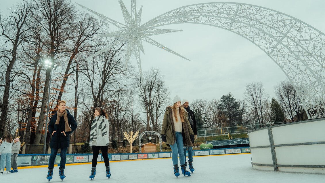 Ein Eislaufplatz mit mehreren Personen, die auf dem Eis entspannt skaten. Im Hintergrund sind Bäume und eine dekorative Sternenskulptur der Winterwelt Graz zu sehen. | © Region Graz-studio draussen