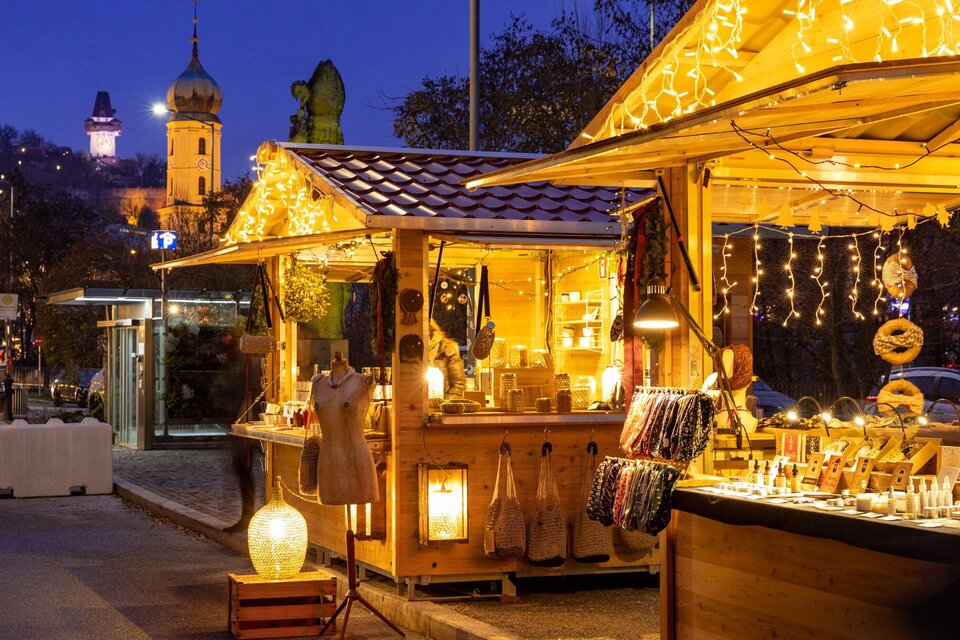 Ein festlich beleuchteter Weihnachtsmarkt mit Holzständen. Im Hintergrund sind Lichter und eine Kirchturmansicht zu sehen. | © Graz Tourismus - Harry Schiffer