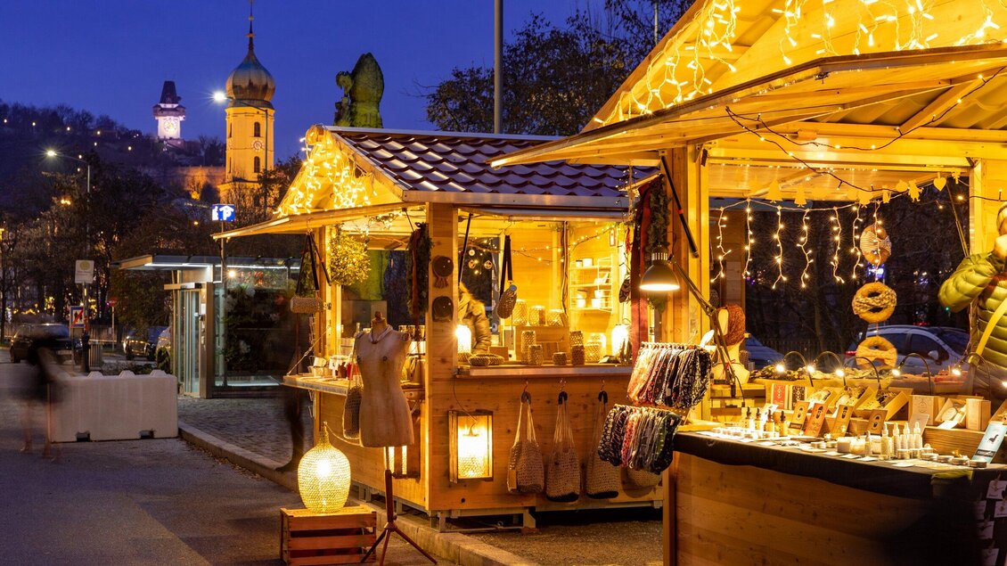 Ein festlich beleuchteter Weihnachtsmarkt mit Holzständen. Im Hintergrund sind Lichter und eine Kirchturmansicht zu sehen. | © Graz Tourismus - Harry Schiffer
