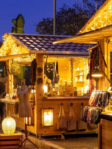 Ein festlich beleuchteter Weihnachtsmarkt mit Holzständen. Im Hintergrund sind Lichter und eine Kirchturmansicht zu sehen. | © Graz Tourismus - Harry Schiffer