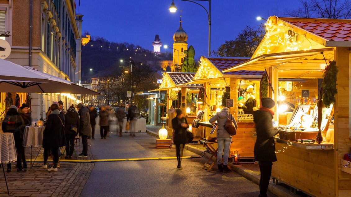 Ein festlicher Weihnachtsmarkt bei Nacht mit beleuchteten Ständen. Menschen schlendern vorbei und genießen die festliche Atmosphäre. | © Graz Tourismus - Harry Schiffer