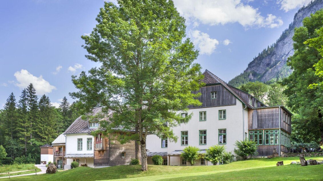 Ein schönes Gebäude umgeben von Bäumen und Wiesen. Im Hintergrund sind Berge zu sehen. | © JUFA Hotel Grundlsee