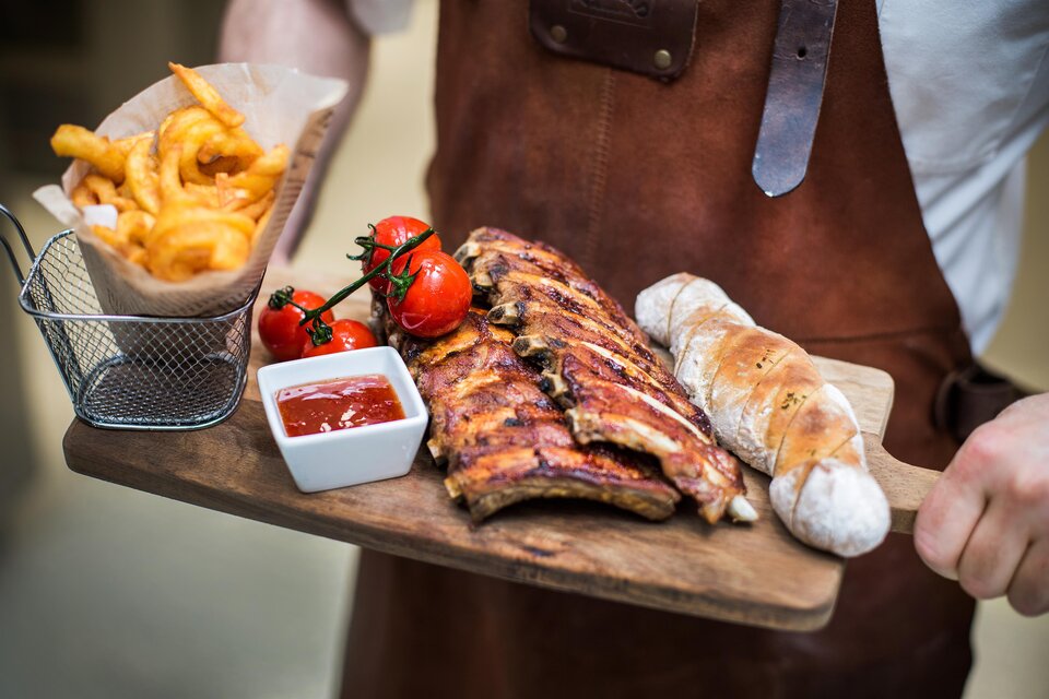 Ein Holzbrett mit gegrillten Rippchen, frischen Tomaten, einem Brötchen und einer Portion Pommes. Dazu gibt es eine kleine Schale mit Soße. | © Painer Katrin