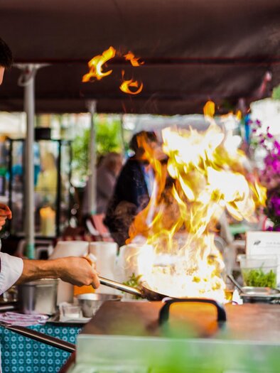 Ein Koch bereitet ein Gericht auf einem Grill vor, während Flammen auflodern. Im Hintergrund sind Gäste und blühende Pflanzen zu sehen. | © @KollarGöbl