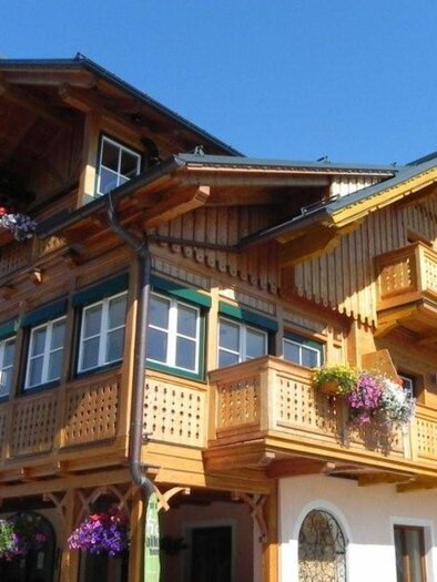 A traditional hotel adorned with flower boxes and wooden balconies is illuminated by the sun. In the background, the sky shines blue. | © Kanzler