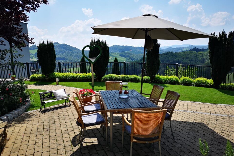 Ein gemütlicher Garten mit einem Esstisch, Stühlen und einem Sonnenschirm. Im Hintergrund sind grüne Hügel und ein blauer Himmel zu sehen. | © TV Südsteiermark
