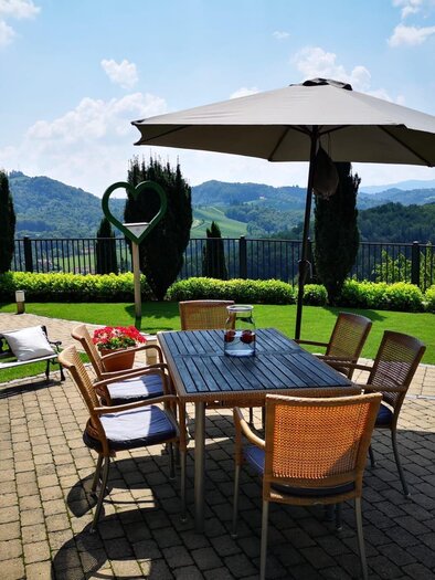 A cozy garden with a dining table, chairs, and a sun umbrella. In the background, there are green hills and a blue sky. | © TV Südsteiermark