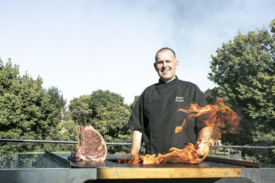 Ein Koch steht mit einem lächelnden Gesicht vor einem Grill, während Flammen hochschlagen. Neben ihm liegt ein großes Stück Fleisch auf dem Grill. | © Vulkanlandhotel Legenstein