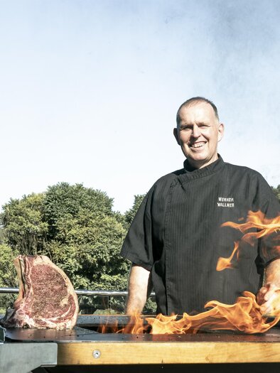 A chef stands with a smiling face in front of a grill, while flames shoot up. Next to him, a large piece of meat lies on the grill. | © Vulkanlandhotel Legenstein