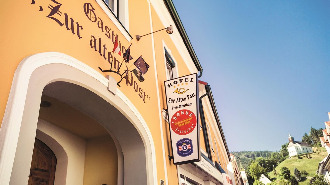 Ein traditionelles Gasthaus mit der Aufschrift „Zur alten Post“. Im Hintergrund sind grüne Hügel und ein blauer Himmel sichtbar. | © Mauthners