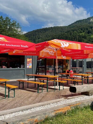 A cozy snack bar with several tables and red umbrellas. In the background, green mountains and a blue sky can be seen. | © Auszeit Ausseerland