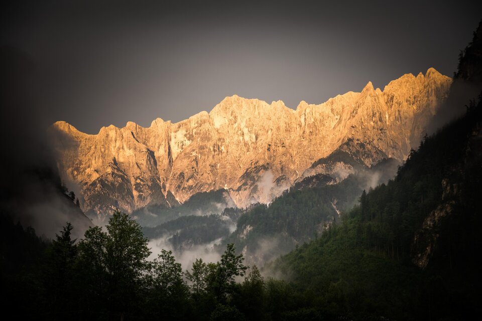 Ein majestätisches Bergpanorama mit Gipfeln, die im Licht der untergehenden Sonne leuchten. Dichte Wälder und Nebel umgeben die Landschaft.