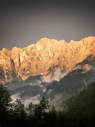 Ein majestätisches Bergpanorama mit Gipfeln, die im Licht der untergehenden Sonne leuchten. Dichte Wälder und Nebel umgeben die Landschaft.