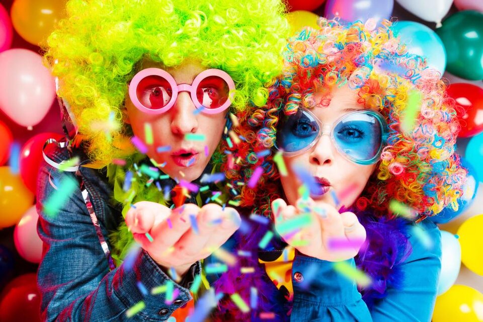 Zwei Personen in bunten Perücken und großen Sonnenbrillen blasen Konfetti in die Luft. Im Hintergrund sind farbige Luftballons zu sehen. | © shutterstock