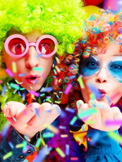 Zwei Personen in bunten Perücken und großen Sonnenbrillen blasen Konfetti in die Luft. Im Hintergrund sind farbige Luftballons zu sehen. | © shutterstock
