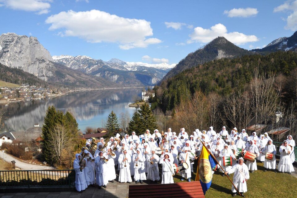 Die Gruppe der traditionellen "Grundlseer Trommelweiber" in ihren weißen Gewändern und Masken steht vor der malerischen Berglandschaft von Grundlsee, mit dem markanten Berg "Backenstein" auf der linken Seite und dem Berg "Ressen" auf der rechten Seite. Die Trommler halten Fahnen und Musikinstrumente, während die Sonne scheint. | © AmonHarry