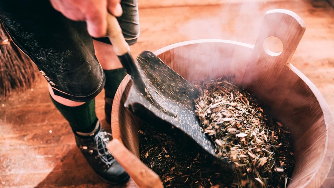 Eine Person rührt mit einer Schaufel in einem Holzbehälter mit dampfenden Inhalten. Die Szene spielt sich in einem rustikalen Innenraum ab. | © Armin Walcher