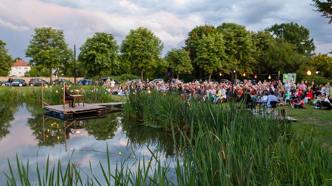 Ein unterhaltsames Freiluft-Event am Wasser mit einer großen Menschenmenge. Bäume umgeben die Szene, während eine Person auf einer Bühne auftritt. | © ORF-Regine Schöttl