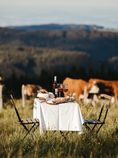 Ein romantisches Picknick auf einer Wiese mit Blick auf sanfte Hügel. Der Tisch ist gedeckt mit Wein, Brot und Käse, während Kühe im Hintergrund grasen. | ©  Achromaticphotography