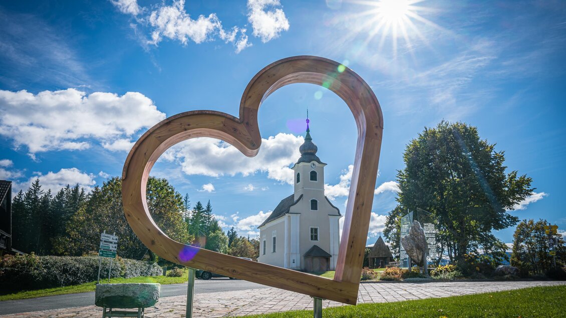 Eine herzförmige Holzskulptur umrahmt eine kleine Kirche im Freien. Der Himmel ist klar mit einigen Wolken und strahlendem Sonnenschein. | © Christian Freydl