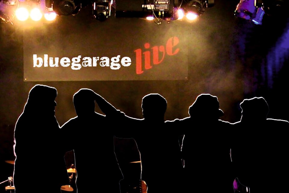 Eine Gruppe von Menschen steht silhouettenartig vor einer Bühne mit bunten Lichtern. Im Hintergrund ist ein Banner mit der Aufschrift "bluegarage live" zu sehen. | © Bluegarage