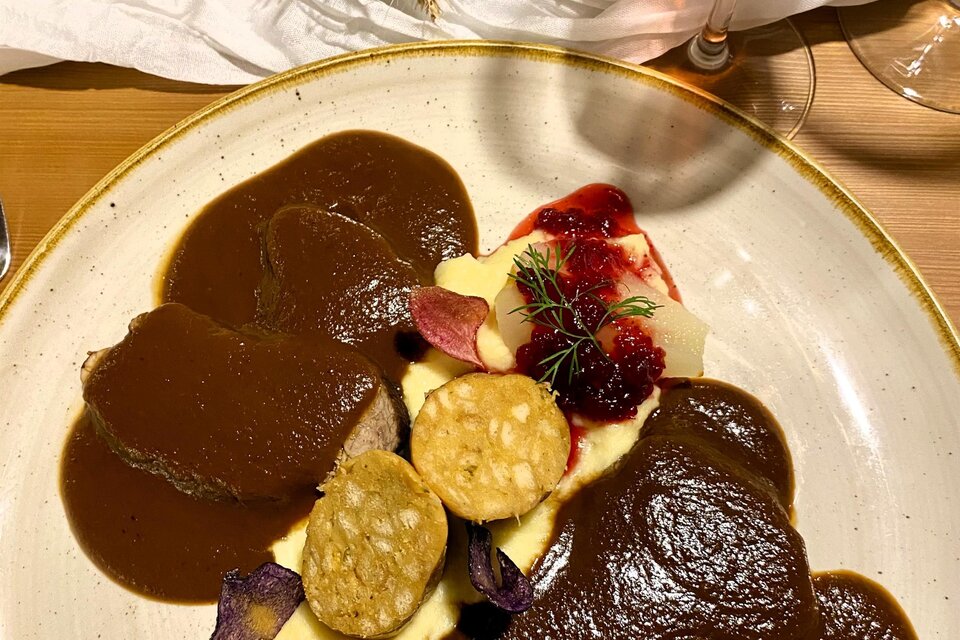 Ein Teller mit zartem Fleisch in einer dunklen Soße, serviert mit Püree und dekorativen Beilagen. Im Hintergrund ist ein Getränk und eine Blumenarrangement zu sehen. | © Trieb