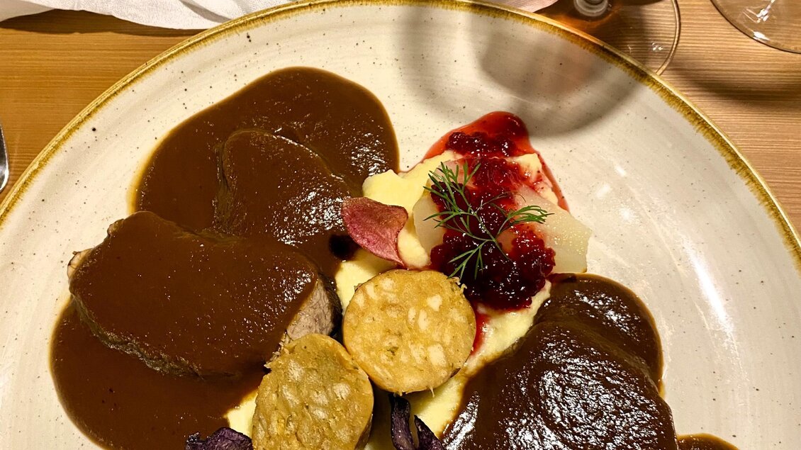 Ein Teller mit zartem Fleisch in einer dunklen Soße, serviert mit Püree und dekorativen Beilagen. Im Hintergrund ist ein Getränk und eine Blumenarrangement zu sehen. | © Trieb