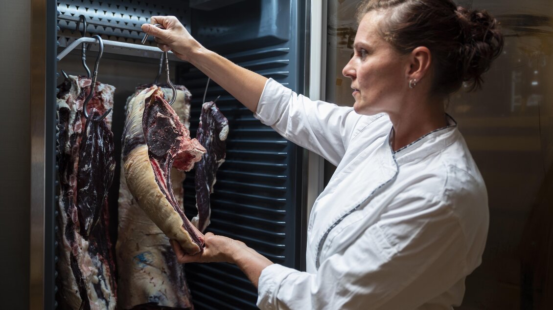 Eine Köchin untersucht Fleischstücke in einem Kühlschrank. Sie trägt eine weiße Kochjacke und schaut konzentriert auf das Fleisch. | © Bergmann B.