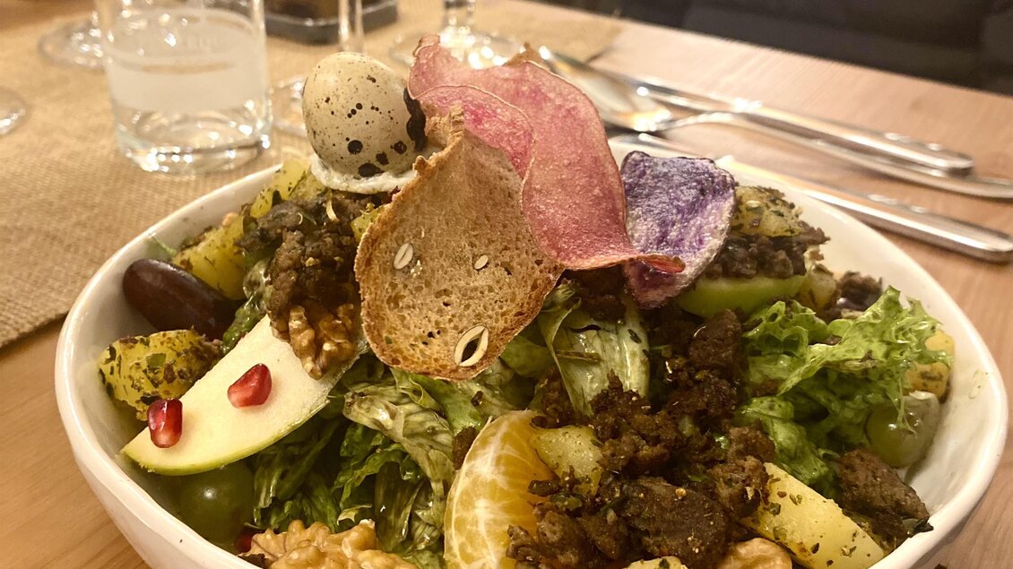 Ein bunter Salat mit frischem Gemüse, Nüssen und essbaren Dekorationen. Er ist in einer weißen Schüssel serviert und umgeben von Gläsern und Tischgedecken. | © Trieb