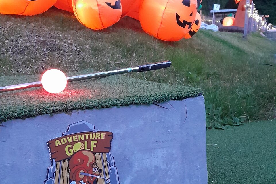 Eine bunte Minigolfanlage mit leuchtenden Kürbissen und einem Schild, das die Nummer eins anzeigt. Die Umgebung ist grün und von Bäumen umgeben.