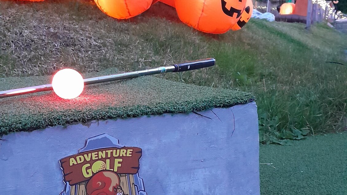 Eine bunte Minigolfanlage mit leuchtenden Kürbissen und einem Schild, das die Nummer eins anzeigt. Die Umgebung ist grün und von Bäumen umgeben.