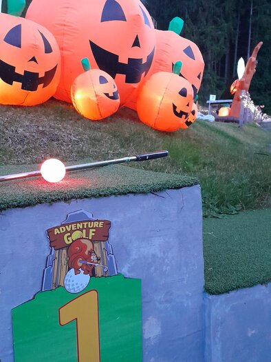 Eine bunte Minigolfanlage mit leuchtenden Kürbissen und einem Schild, das die Nummer eins anzeigt. Die Umgebung ist grün und von Bäumen umgeben.