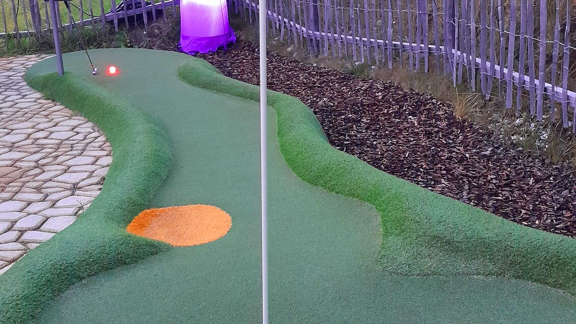 Eine Minigolfbahn mit einem Loch und einer Fahne. Im Hintergrund steht eine leuchtende Figur.