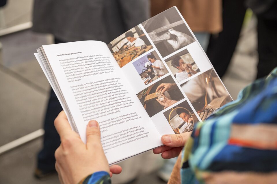 Eine Person hält einen geöffneten Ausstellungskatalog mit Texten und Fotos zum Handwerk auf der Murinsel in Graz. | © Bernd Grosseck