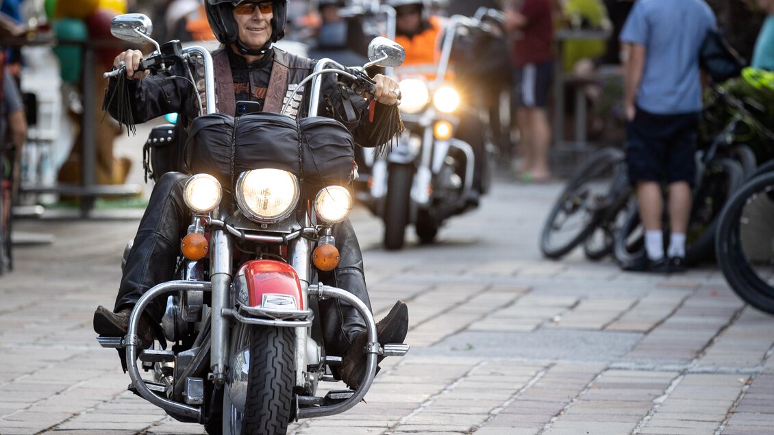 Ein Motorradfahrer fährt auf einer Straße, umgeben von Zuschauern. Im Hintergrund sind weitere Motorräder und Menschen zu sehen. | © Harald Steiner