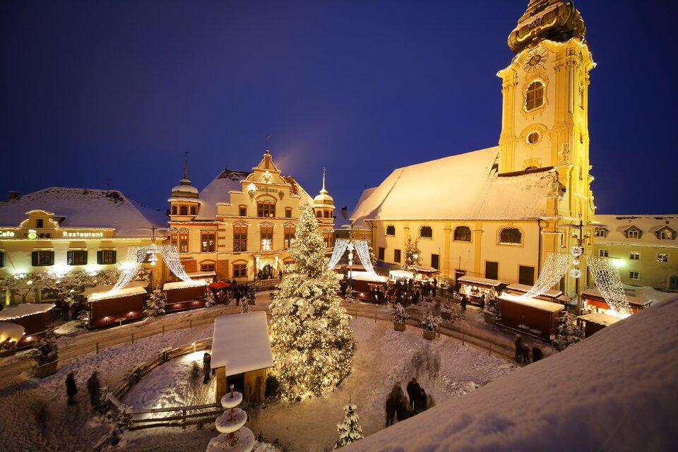 Ein festlicher Weihnachtsmarkt mit festlich beleuchteten Ständen und einem großen, geschmückten Baum. Der Platz ist von schneebedeckten Gebäuden und einer Kirche umgeben, alles unter einem klaren Nachthimmel. | © Bernhard Bergmann