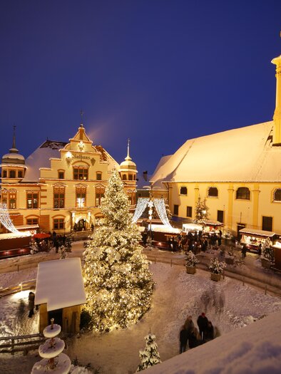 A festive Christmas market with festively lit stalls and a large decorated tree. The square is surrounded by snow-covered buildings and a church, all under a clear night sky. | © Bernhard Bergmann