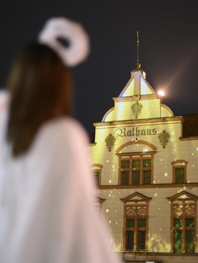 A person in an angel costume stands in front of a festively lit building. The sky is dark, and the lights radiate a Christmas atmosphere. | © Tourismus Oststeiermark